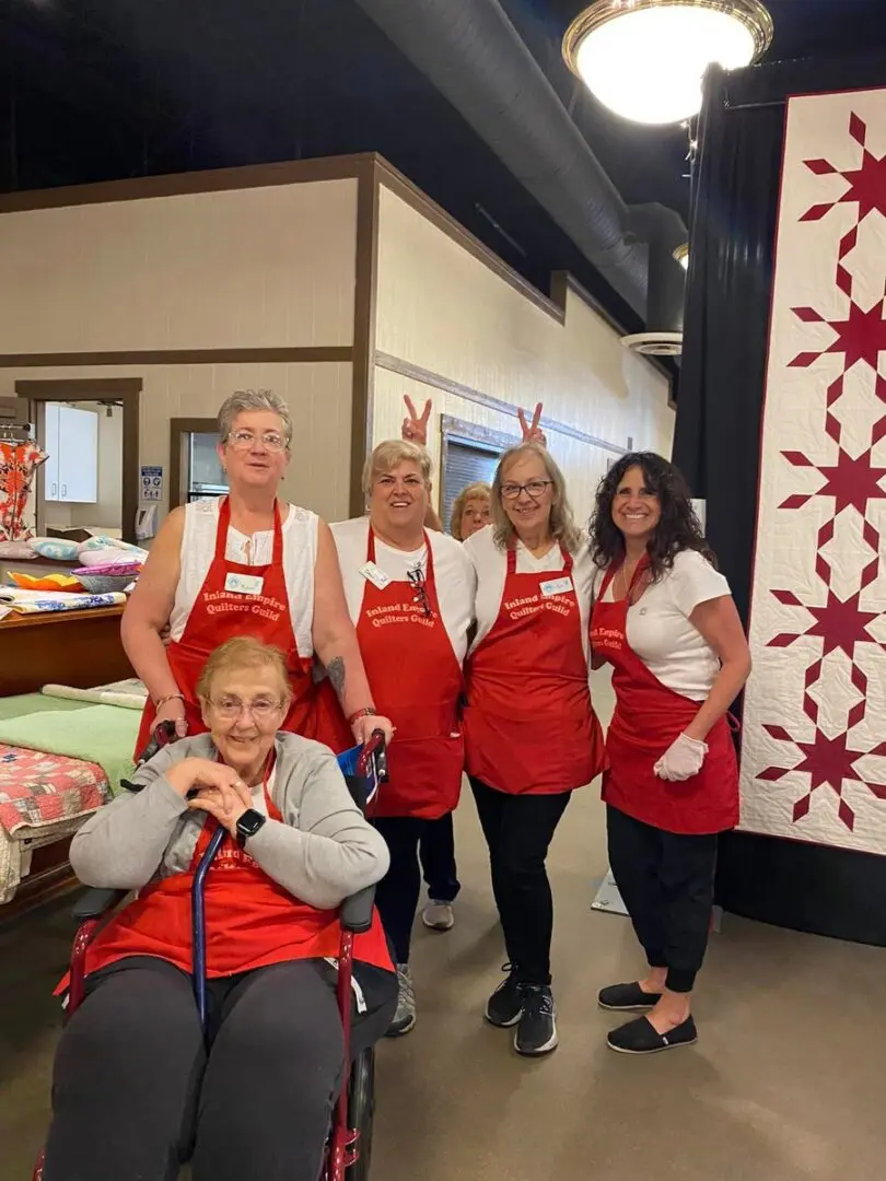 Group of women in red aprons posing for a photo in front of a quilt