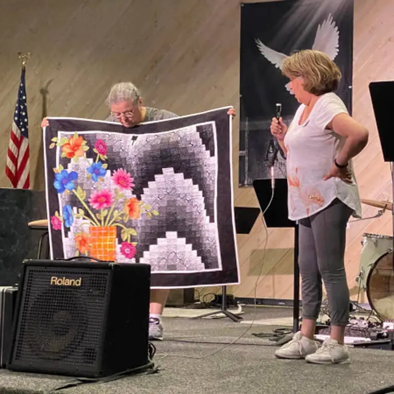 Woman standing on a stage, holding a microphone in her hand and a quilt in front of her