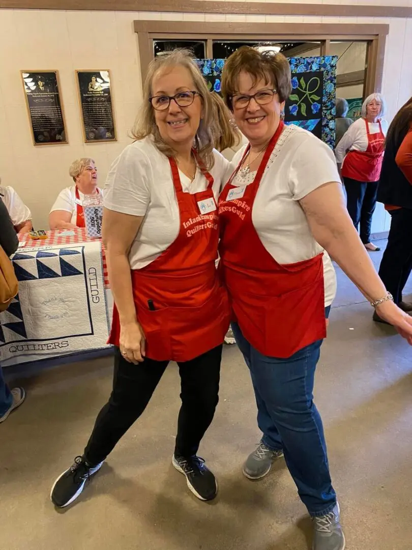 Two women in red aprons standing next to each other, Behind them is a table