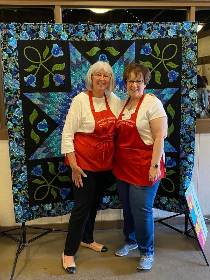 Two women in red aprons standing next to each other in front of a quilt
