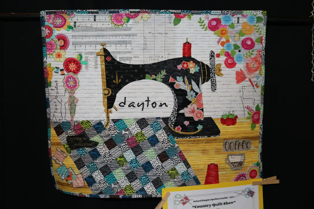 Quilted wall hanging with a sewing machine on it, hung on a wall with a board and a thread roll