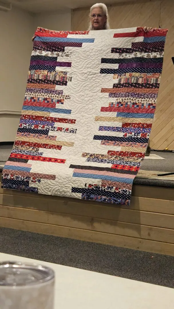 Woman standing in front of a table, holding up a quilt made with a variety of fabrics