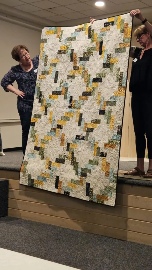 Two women standing on a stage, holding up a quilt that is being displayed