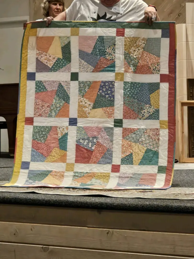 Man and woman standing on a stage, holding up a quilt that is being made by the woman