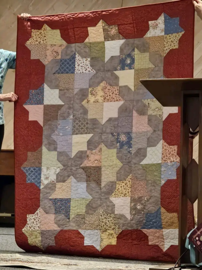 Two people holding up a large quilt in front of a podium, with a wall in the background
