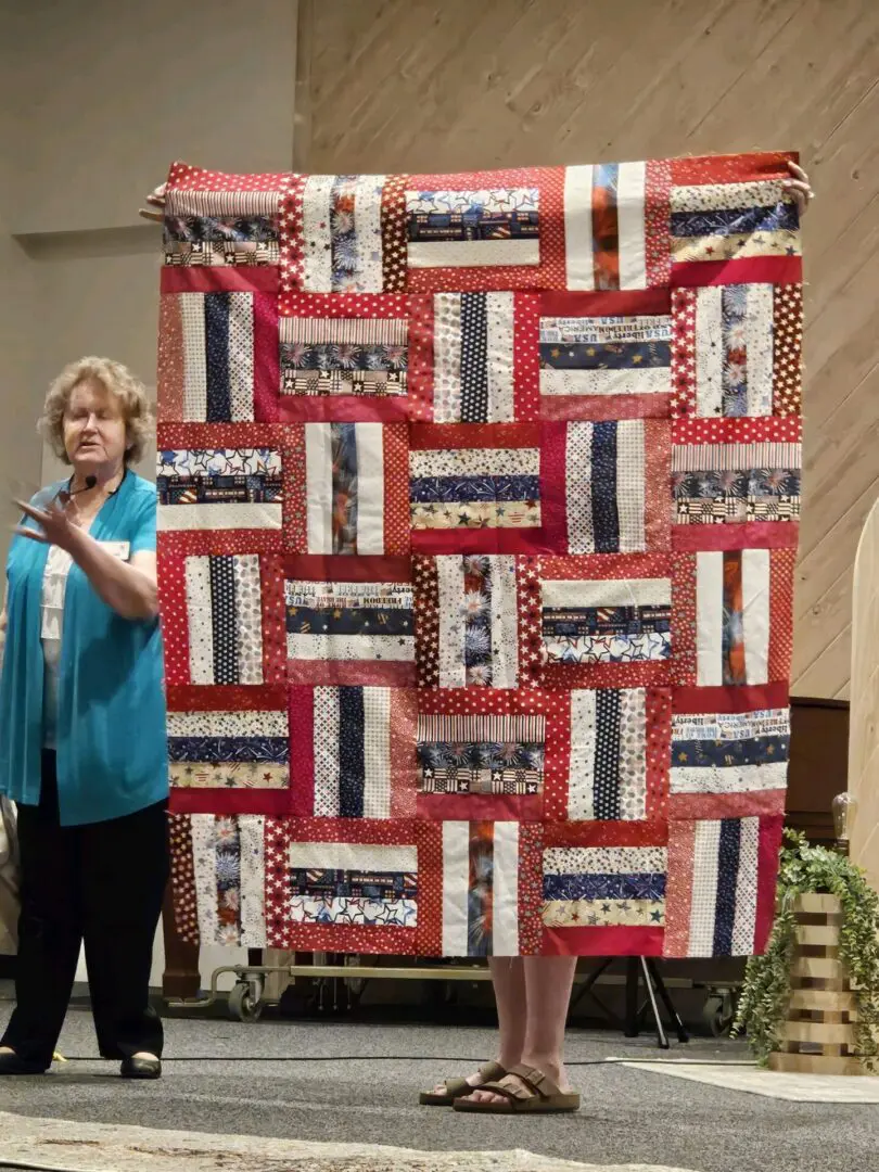Woman standing on a stage, holding up a large quilt with a variety of colors and patterns