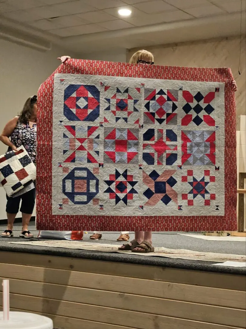 Two women standing on a stage, holding up a quilt with a red, white and blue pattern