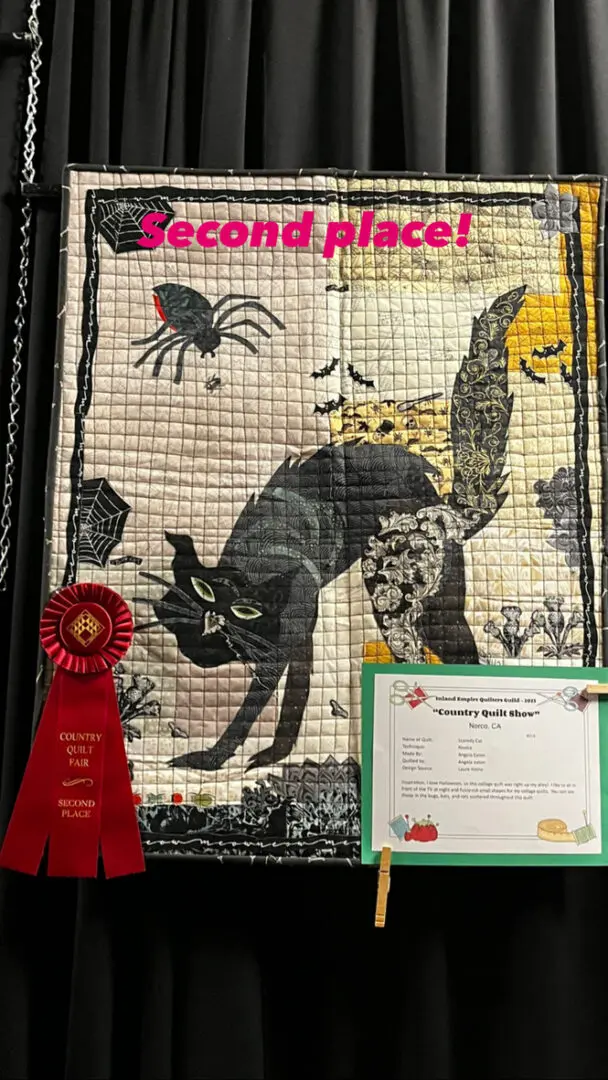 Quilt with a black cat on it, hanging on a wall with a red ribbon