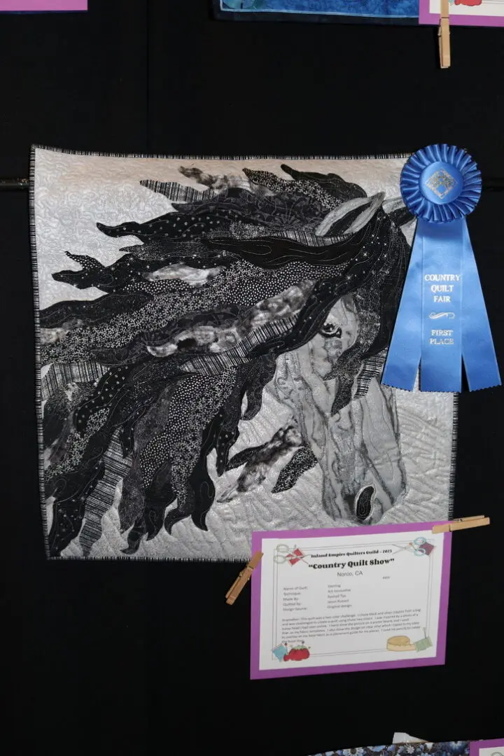 Quilted wall hanging with a horse on it, attached to a black surface with a blue ribbon