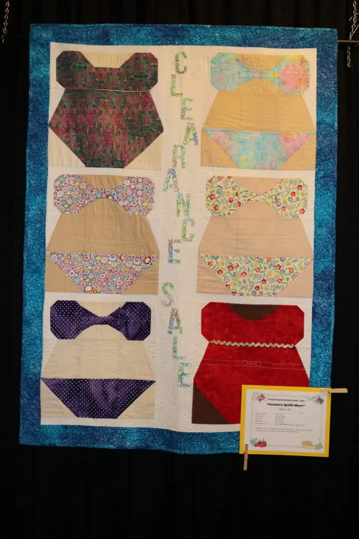 Quilted wall hanging on a black curtain, featuring a variety of colorful clothes and a certificate attached to it