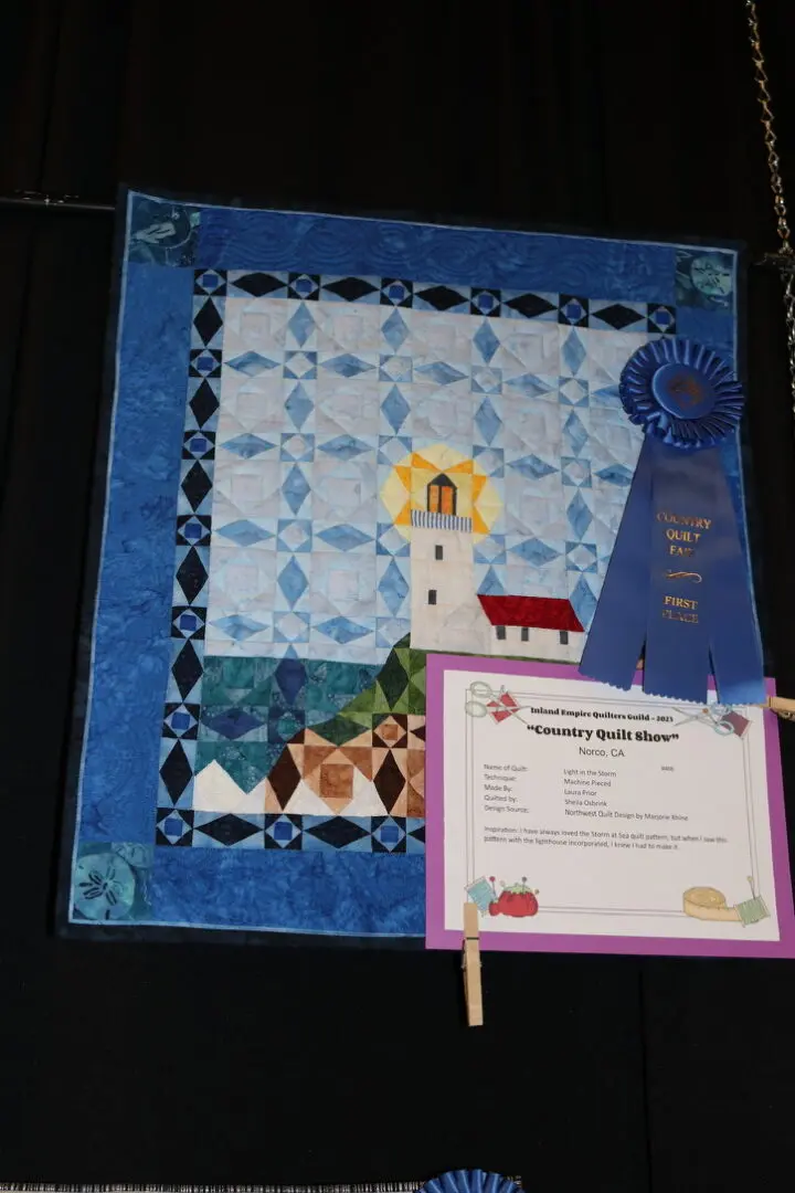 Quilted wall hanging with a blue ribbon on it, attached to a black cloth with a chain