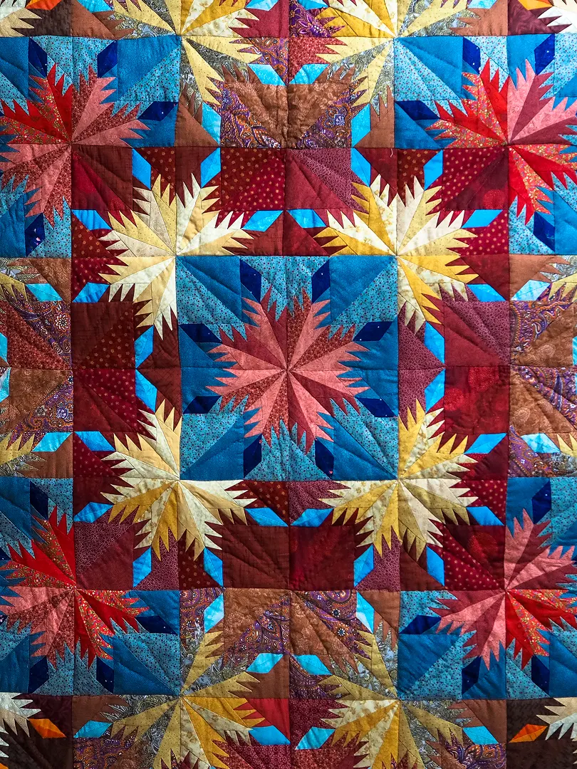 Colorful quilt featuring repeating eight-point starburst patterns in red, gold, turquoise, and deep blue; intricate stitching gives the geometric design a vibrant, festive feel.