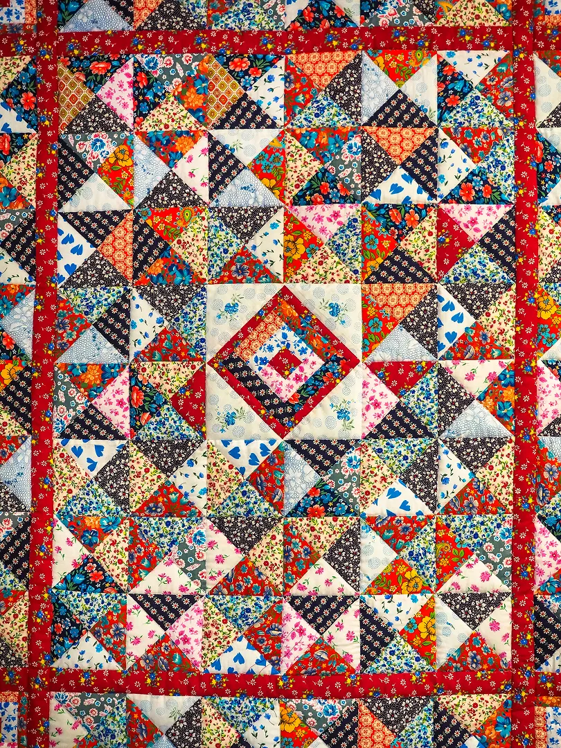 A vibrant patchwork quilt filled with floral prints and geometric triangles, centered on a layered diamond medallion and framed by a bold red border.