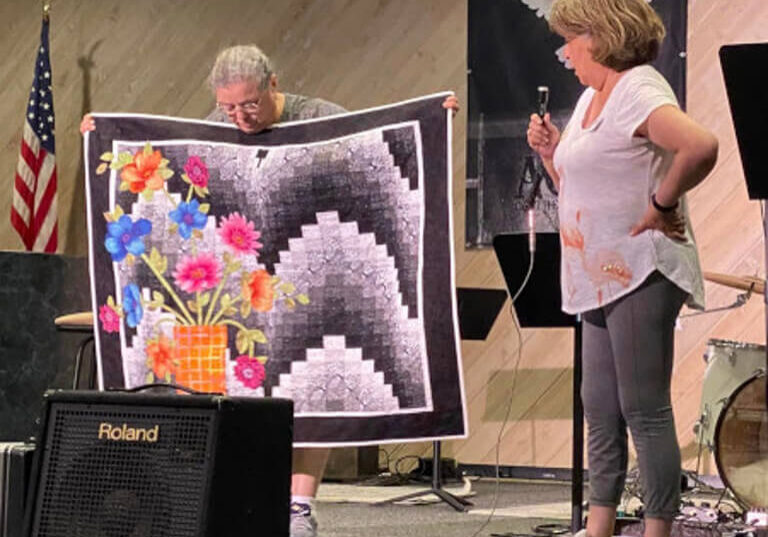 Woman standing on a stage, holding a microphone in her hand and a quilt in front of her