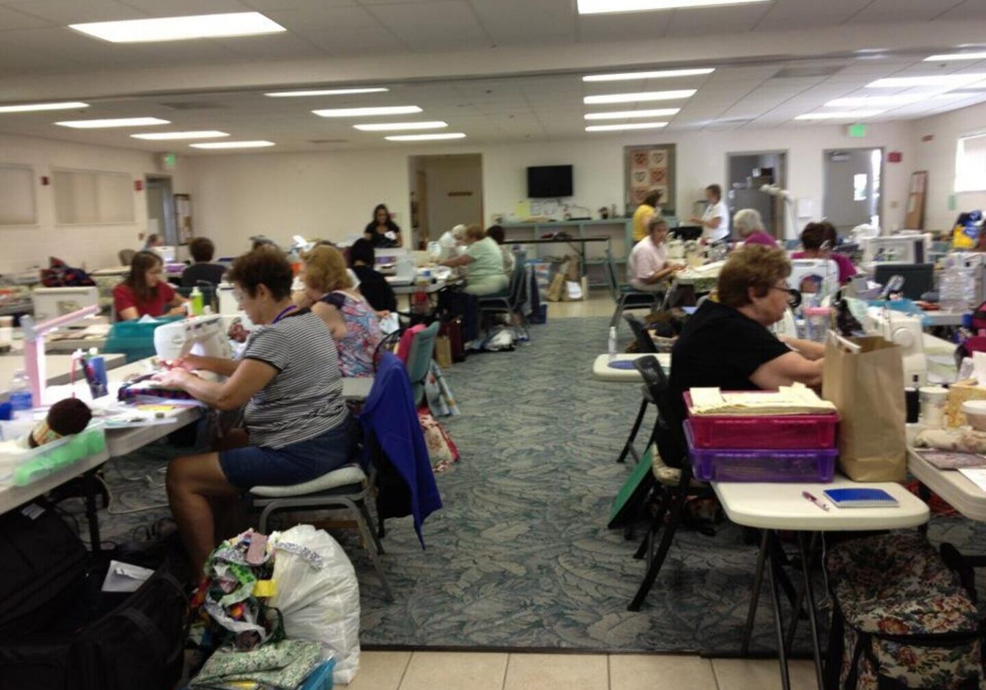Large room filled with lots of people sitting at tables, sewing machines, clothes, bags, and other objects